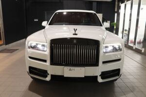 Front view of a white Rolls-Royce SUV in a showroom, with a prominent vertical grille and bright LED headlights.