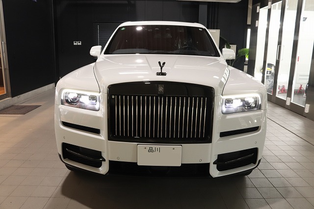Front view of a white Rolls-Royce SUV in a showroom, with a prominent vertical grille and bright LED headlights.