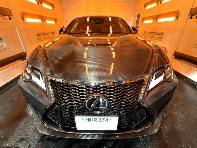 Front view of a Lexus sedan with a carbon fiber hood inside an orange-lit detailing bay; prominent spindle grille and Lexus emblem visible on the bumper.