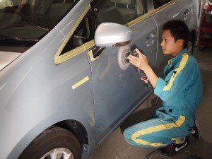 プリウス コンクリートミスト除去 研磨 - トータルカービューティIIC Auto body technician in blue uniform polishing a car door with a buffer in a workshop, masking tape around the window area.