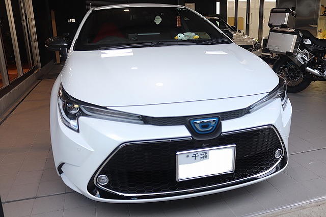 Front view of a white car displayed in a dealership showroom, featuring a black honeycomb grille, angular headlights, and a blurred license plate.
