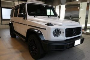 White Mercedes-Benz G-Class SUV parked indoors in a showroom with black wheels and grille detail visible.