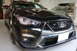 Front view of a black Nissan sedan in a showroom, highlighting the chrome grille and LED headlights at the front corner.