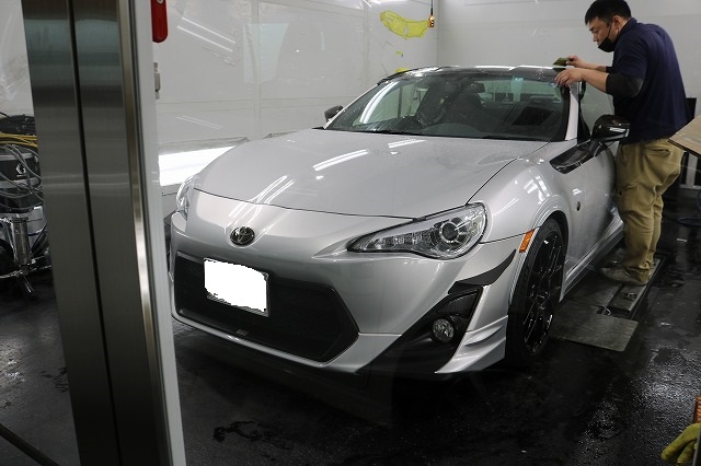 Front three-quarter view of a silver Toyota sports car in a workshop, a technician in a mask wheels at the windshield.