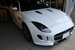 Front three-quarter view of a white Jaguar sports car in a showroom, showcasing the sculpted hood and aggressive front bumper.