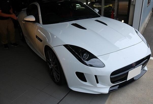 Front three-quarter view of a white Jaguar sports car in a showroom, showcasing the sculpted hood and aggressive front bumper.