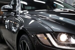 Close-up of a dark gray car's front left quarter, with a lit headlight and reflective hood edge.