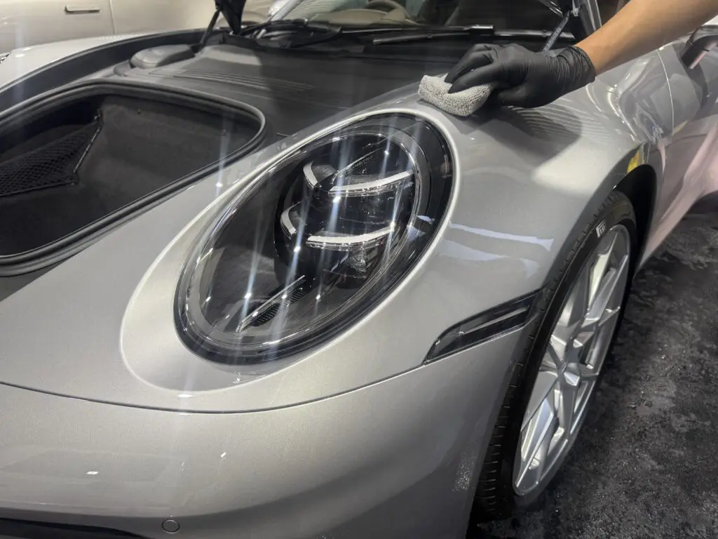 Person wearing black glove polishing a silver car hood around the headlight with a cloth.