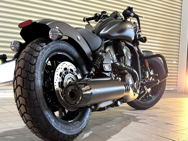 インディアン スカウトボバー リア写真 - トータルカービューティIIC Rear three-quarter view of a matte black Indian motorcycle, showing a large rear tire, disc brake, and dual exhaust.