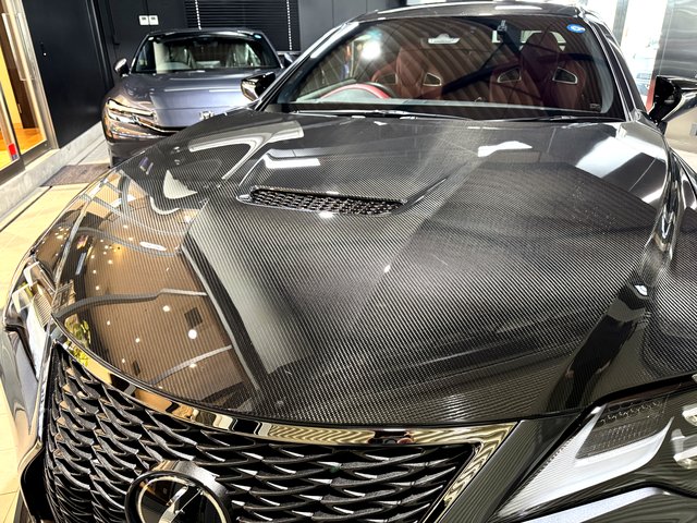 Front view of a black luxury sedan with a carbon fiber hood on display in a showroom, reflections on the hood.
