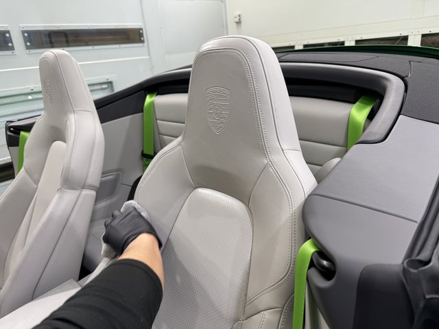 Light gray leather Porsche convertible interior with embossed logo on the headrest; lime green seat belts visible, a gloved hand touching the seat edge.
