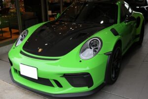 Front view of a lime-green Porsche sports car with a black hood, parked indoors in a showroom; round headlights visible.