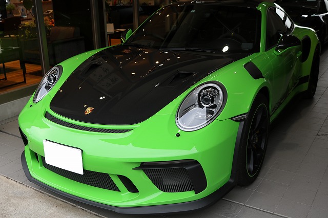 Front view of a lime-green Porsche sports car with a black hood, parked indoors in a showroom; round headlights visible.