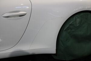 Close-up of a white car's rear quarter panel showing a small dent and scratch near the wheel, with the wheel covered by a green protective cover