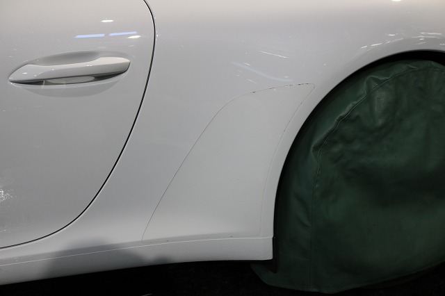 Close-up of a white car's rear quarter panel showing a small dent and scratch near the wheel, with the wheel covered by a green protective cover