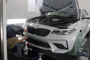 IMG_0524 - トータルカービューティIIC Auto detailer applies treatment to the front bumper of a white BMW with the hood raised in a workshop.