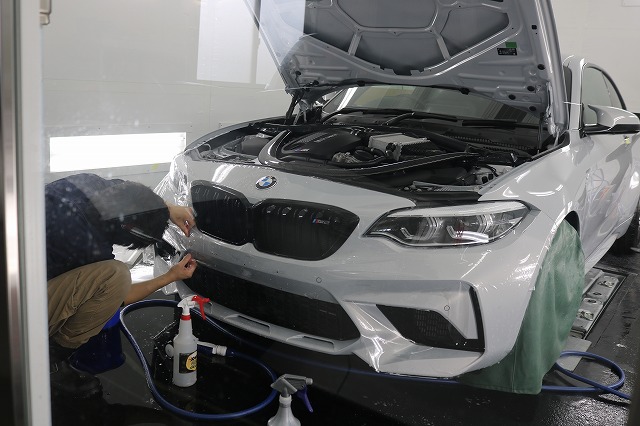 Auto detailer applies treatment to the front bumper of a white BMW with the hood raised in a workshop.