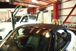 Black convertible parked inside an auto repair shop, with a white car and yellow ladders in the background.